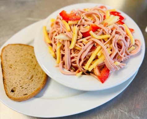 Bunter Nudelsalat mit Schinken, Paprika und einem Stück Brot auf einem weißen Teller.