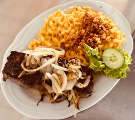 Teller mit gegrilltem Steak, Käse-Nudeln, Zwiebeln, Salat und Gurkenscheibe.